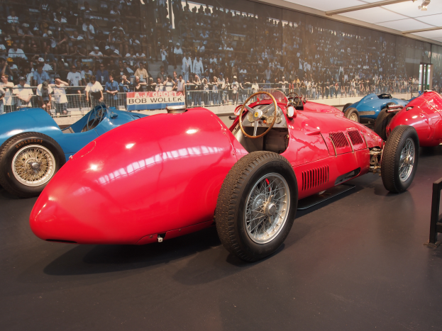 Ein rotes Rennauto in einem Museum ausgestellt, umgeben von anderen Autos, mit einem Stand auf der rechten Seite, einer Werbetafel im Hintergrund und Lichtern auf dem Dach, die eine helle Atmosphäre schaffen.