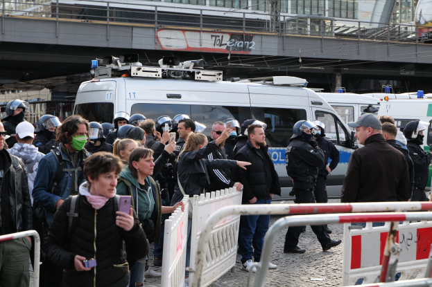 Gruppe von Menschen vor Polizeiwagen mit Absperrungen im Vordergrund und einer Brücke im Hintergrund bei einer Demonstration in Berlin, Deutschland.