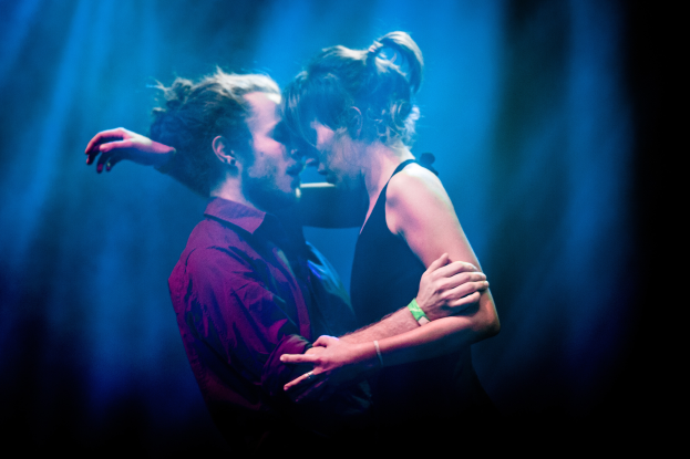 Ein Mann und eine Frau umarmen und küssen sich auf einer Bühne, während einige Lichter den Hintergrund beleuchten.