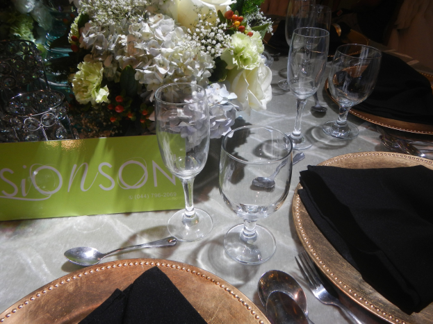 Formaler Esstisch mit goldenen Untersetzern, schwarzen Servietten, Besteck, Gläsern und einem Blumenstrauß, mit einer Tafel mit Text.