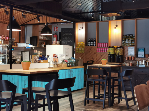 Innenraum eines Restaurants mit Holztischen, Stühlen, einer Topfpflanze auf einem Tisch, einer Schiefertafelmenü, einem Kühlschrank, einem Feuerlöscher, Regalen mit Flaschen, Schränken mit Gegenständen, einem gerahmten Foto an der Wand und Deckenleuchten.