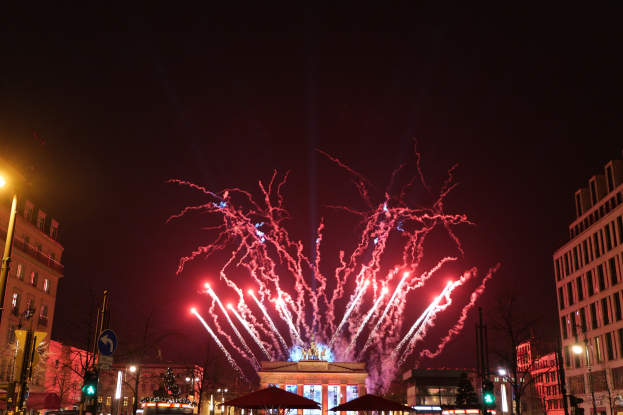 Eine belebte Stadtstraße in Berlin am Neujahrstag, voller Menschen, Fahrzeugen und Gebäuden, beleuchtet von Lichtern und Feuerwerken am Himmel.