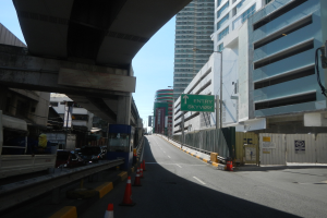 Stadtstraße mit Verkehrsleitkegeln am Straßenrand, Fahrzeuge auf der Straße, eine Brücke links, Gebäude mit Namensschildern auf beiden Seiten, Laternenmasten und ein Himmel im Hintergrund.