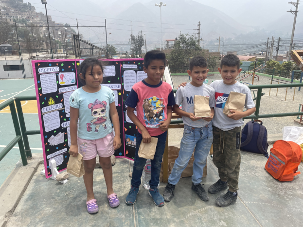 Gruppe von Kindern auf einem Basketballfeld, die Papiertüten halten, mit Tüten und Gegenständen rechts und einer Tafel mit Text links, vor dem Hintergrund von Zäunen, Pfosten, Straßenlaternen, Strommasten, Bäumen, Gebäuden, Hügeln und Himmel.