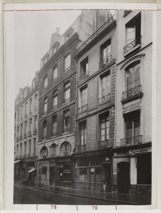 Schwarzes und wei├čes Foto einer Stadtstra├če mit Geb├Ąuden mit Fenstern, Gel├Ąndern und Namensschildern und einer Stra├če unten.