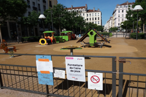 Umzäunter Spielplatz auf einem städtischen Platz mit Schildern am Zaun, Bäumen, Laternenmasten und Gebäuden im Hintergrund bei sichtbarem Himmel.