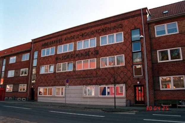 Ein großes Backsteingebäude mit vielen Fenstern, das als Hauptquartier der deutschen Bundespolizei dient, befindet sich an einer Straße mit einem Gehweg, einem Baum, einer Bank, einem Laternenpfahl und einem roten Kasten, mit dem Himmel im Hintergrund.