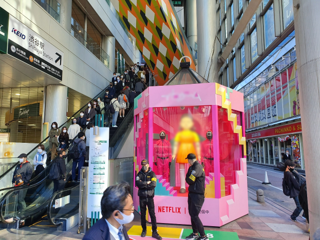 Eine Gruppe von Menschen steht vor einem Gebäude mit Glaswänden, Säulen und Texttafeln, einige tragen Masken, mit einer Rolltreppe im Blick; eine pinkfarbene Box in der Mitte zeigt Schaufensterpuppen und trägt die Aufschrift "Netflix's new Netflix booth in Tokyo."
