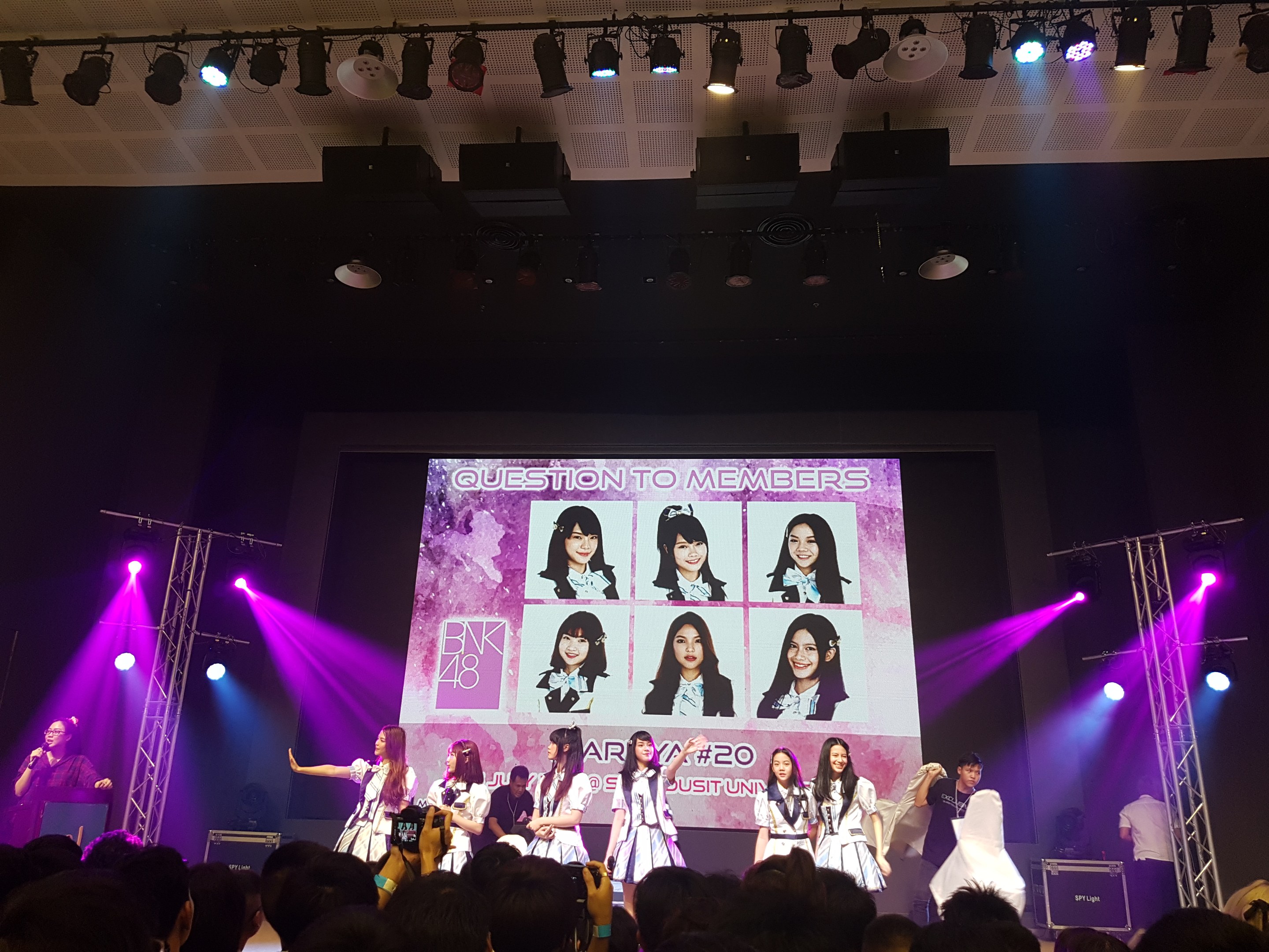 Eine Gruppe von Menschen auf einer hell erleuchteten Bühne, die vor einem Publikum spielt, wobei einer ein Mikrofon hält und ein anderer ein Instrument spielt, während ein großer Bildschirm Bilder der K-Pop-Gruppe BK48 anzeigt.