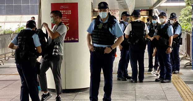 Eine Gruppe von Polizisten in Uniform und Mund-Nasen-Schutz steht vor einem Gebäude, einige halten Kameras, mit einem Banner, Geländern, Pflanzen und Deckenleuchten im Hintergrund.