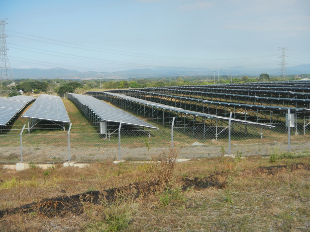 Ein Feld voller Solarpanels, umgeben von einem Zaun, mit Gras und Pflanzen im Vordergrund, Bäumen und Übertragungstürmen im Hintergrund und Hügeln unter einem klaren blauen Himmel.