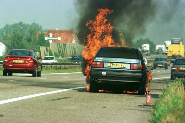 Ein Auto in Flammen neben einer Straße mit anderen Fahrzeugen, Bäumen, Gebäuden und einem blauen Himmel im Hintergrund, mit Gras und einem Feuerlöscher auf der rechten Seite.