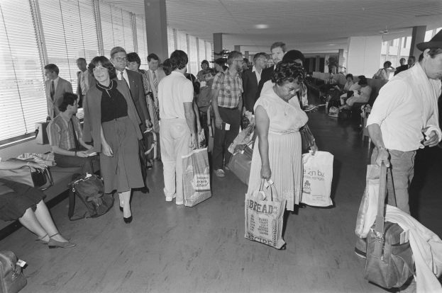 Ein Schwarz-Weiß-Bild von Reisenden in einem Flughafen, einige gehen mit Gepäck, andere sitzen auf Bänken oder stehen, mit einem Mann und einer Frau, die Taschen halten, in der Mitte und Fensterjalousien links.