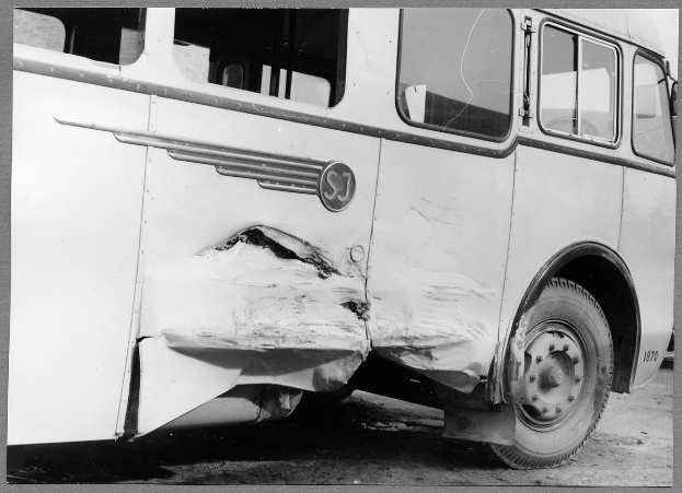 Schwarzes und weißes Foto eines beschädigten Busses am Straßenrand mit einer eingedrückten Front, zersplitterten Fenstern und einer offenen Fahrertür, die den Innenraum zeigt.