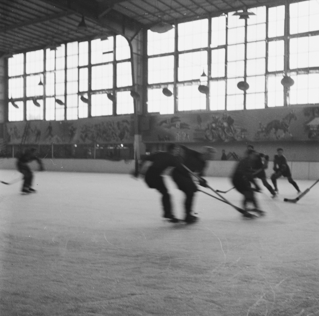 Schwarzes und weißes Foto von Menschen, die Hockey auf einem Eisplatz spielen, mit einer bemalten Wand und Fenstern im Hintergrund.