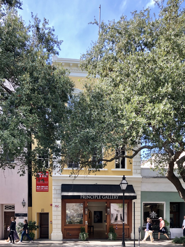 Gruppe von Menschen, die auf einem Gehweg vor der Principle Gallery in Charleston, SC, mit Topfpflanzen, einer Straßenlaterne, einem Schild, einer Fahne, einem Fahnenmast und einem bewölkten Himmel gehen.