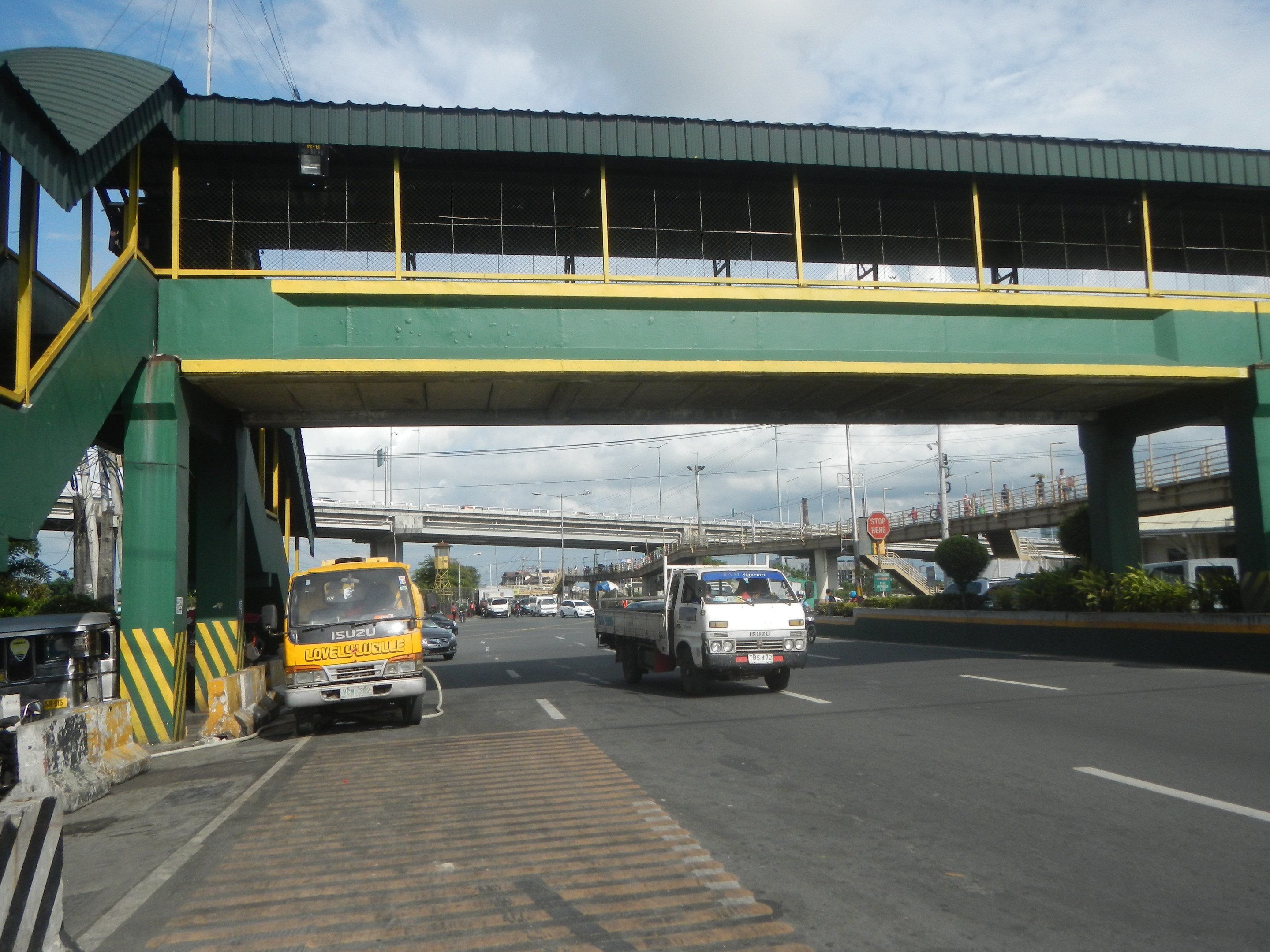Eine Mautstelle mit Fahrzeugen auf einer Straße darunter, eine Brücke mit Geländern und Pfeilern, Laternenmäste, Schilder an Mästen, Bäume und ein bewölktes Himmel im Hintergrund, mit einem Lkw, der auf der Straße neben der Brücke fährt.