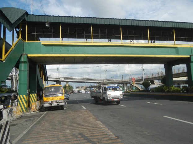 Eine Mautstelle mit Fahrzeugen auf einer Straße darunter, eine Brücke mit Geländern und Pfeilern, Laternenmäste, Schilder an Mästen, Bäume und ein bewölktes Himmel im Hintergrund, mit einem Lkw, der auf der Straße neben der Brücke fährt.