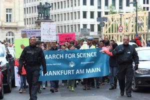 Eine Gruppe von Menschen marschiert mit einer "March for Science Frankfurt am Main"-Fahne die Straße entlang, Autos, Gebäude, Statuen, Laternenpfähle, Schilder und Bäume im Hintergrund.