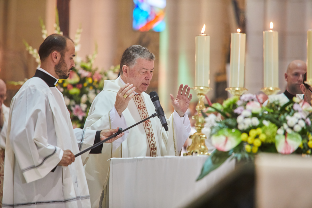 Der Bischof der Kirche des Heiligen Grabes steht an einem Rednerpult mit einem Mikrofon und spricht zu einer Gruppe von Menschen, mit einem Tisch mit Kerzen und Blumen zu seiner Rechten und Blumensträußen und einem Bleiglasfenster im Hintergrund.