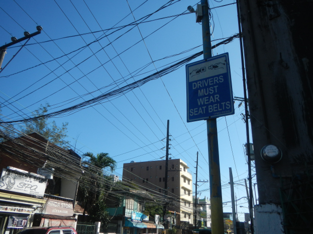 Eine Stadtstraße mit Autos, Strommasten mit Drähten, Gebäuden, Bäumen und Namensschildern, mit einem Schild an einem Mast, auf dem steht "Fahrer müssen Sicherheitsgurte tragen" und der Himmel im Hintergrund.