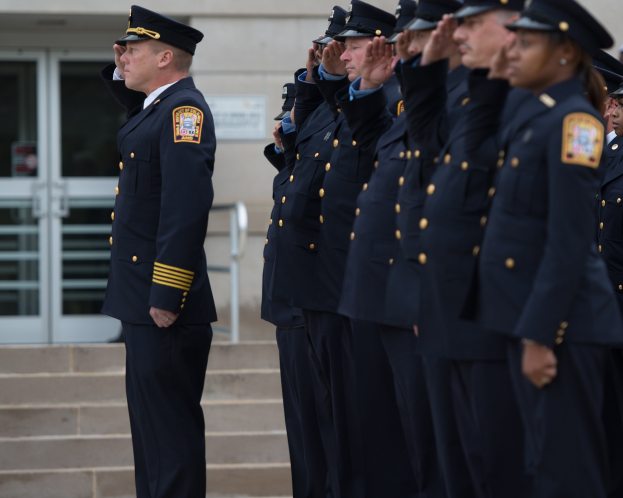 Eine Gruppe von Polizeibeamten in Uniform und Mützen, die in Formation salutieren vor einem Gebäude mit Glastüren und Treppen.