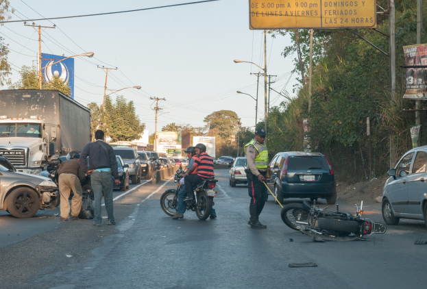 Eine Gruppe von Menschen steht um ein verunglücktes Motorrad auf der Stra├če mit Fahrzeugen, B├Ąumen und Strommasten im Hintergrund.