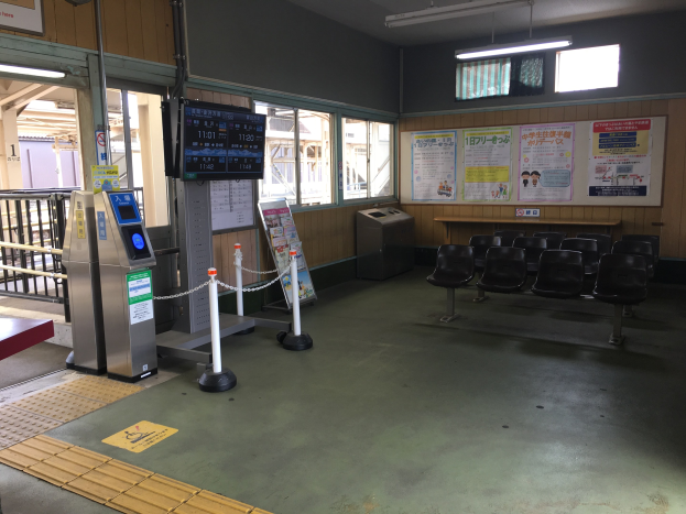 Ein belebter Bahnhofseingang mit Sitzgelegenheiten, einem Informationsschalter, Ticketautomaten, Pfosten mit Ketten, Informationsanzeigen, Wandplakaten, Deckenleuchten und Geländern.