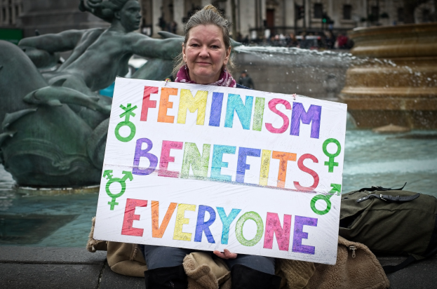Eine Frau sitzt in der Nähe eines Brunnens und hält ein Schild mit der Aufschrift "Feminismus nützt allen" sowie eine Tasche neben sich, während unscharfe Gestalten, eine Statue und ein Gebäude im Hintergrund zu sehen sind.