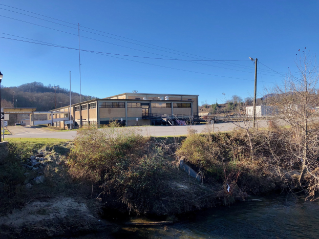 Ein Fluss fließt durch ein grünes Feld neben einem Gebäude, umgeben von Bäumen, Pflanzen, Gras, Steinen, Straßenmasten, Straßenlaternen, Strommasten, Stromkabeln, Hügeln und einem klaren blauen Himmel, mit dem Gebäude als Standort für eine Wasseraufbereitungsanlage vorgeschlagen.