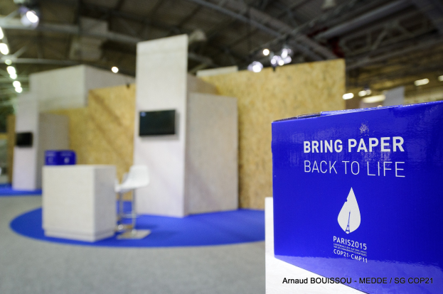 Ein Messestand mit einem blauen und weißen 'Bring Paper Back to Life'-Schild auf der rechten Seite, der undeutliche Stühle, Tische, einen wandmontierten Fernseher und Deckenleuchten im Hintergrund zeigt.