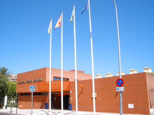 Großes Gebäude mit spanischen Nationalpolizei-Flaggen, umgeben von Straßeninfrastruktur und Bäumen unter einem klaren blauen Himmel.
