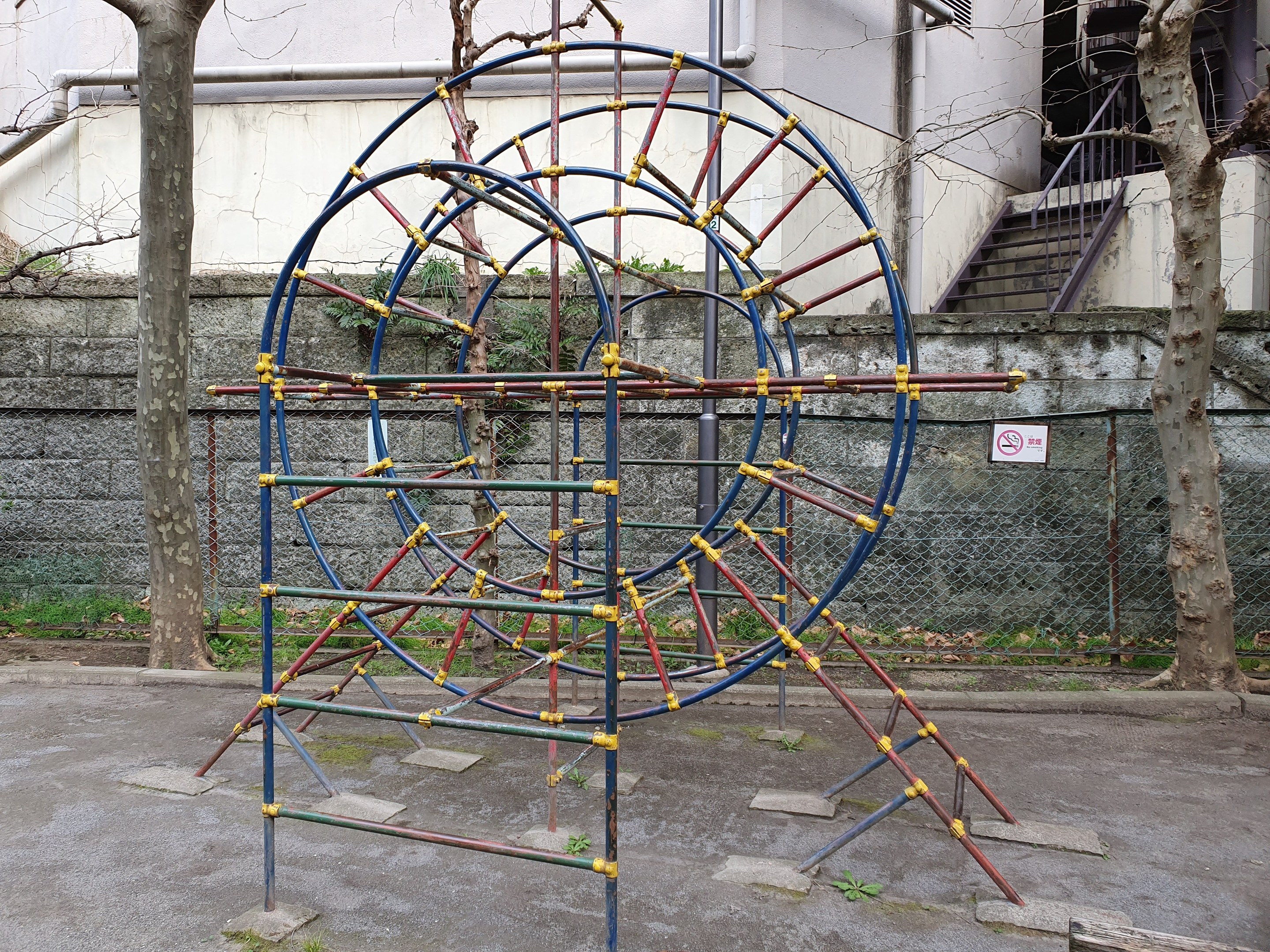 Spielplatz mit großem Metallklettergerüst, umgeben von Bäumen und einer Maschendrahtzaunwand, mit Treppen mit Geländern, die zu einem Gebäude mit Fenstern und Rohren führen, einem Wegweiser und Grasboden.