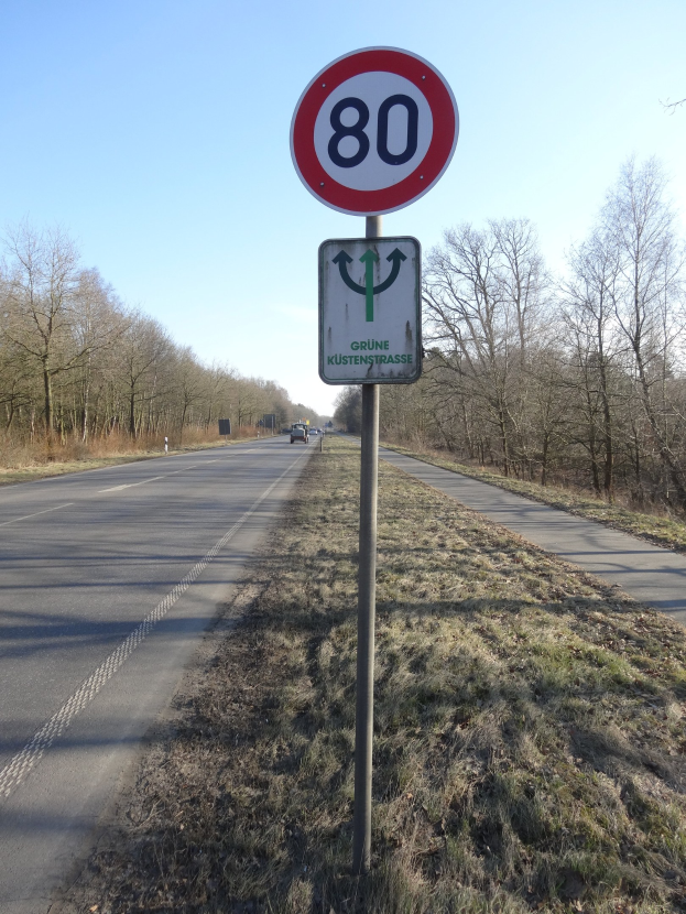 Geschwindigkeitsbegrenzungsschild am Rande einer Straße mit vorbeifahrenden Fahrzeugen, Gras, Bäumen und einem klaren blauen Himmel im Hintergrund.