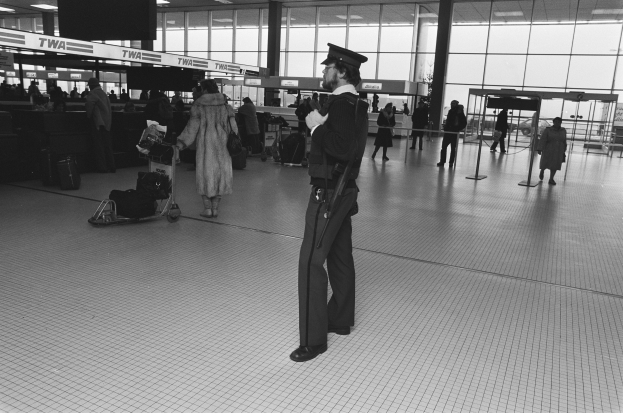 Polizeibeamter in Uniform mit Pistole in einem Flughafen-Terminal umgeben von Menschen und Gepäckstücken, mit Gepäckwagen, Deckenleuchten, Pfosten, Texttafeln und Glaswänden im Hintergrund.