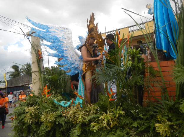 Eine Gruppe von Menschen geht eine Straße entlang neben einem schwimmenden Gerüst, das mit Pflanzen und Blumen geschmückt ist, mit kostümierten Personen auf dem Gerüst, Gebäuden, Bäumen, Strommasten mit Drähten und einem bewölkten Himmel im Hintergrund.