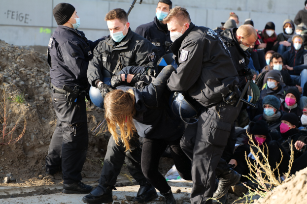 Gruppe maskierter Menschen in Kappen, einige mit Waffen, die eine Frau umgeben, die von der Polizei eskortiert wird, mit Pflanzen, Steinen und einer Wand mit Protesttext im Hintergrund.