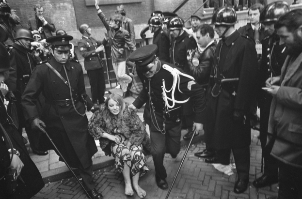 Schwarz-weißes Bild einer Frau, die von der Polizei eskortiert wird, vor einer Menge mit Helmen und Stöcken, mit einem Gebäude mit Treppe und Geländer im Hintergrund.