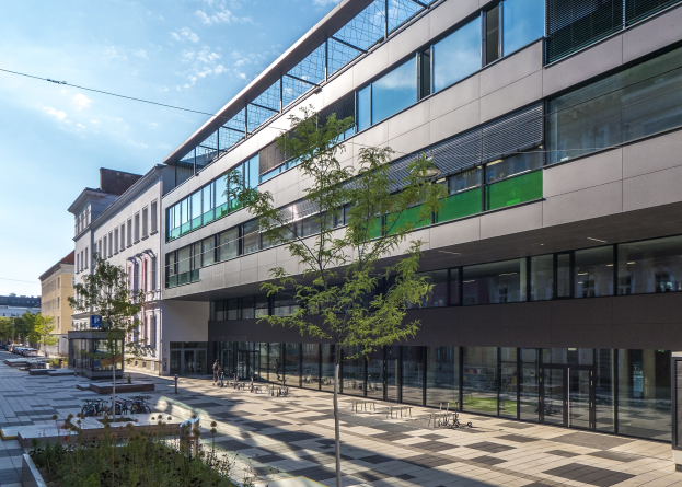Großes modernes Gebäude mit zahlreichen Fenstern, umgeben von Bäumen, Pflanzen, Fahrzeugen und Menschen unter einem bewölkten Himmel, identifiziert als neue deutsche Technologie-Institut-Zentrale in Berlin.
