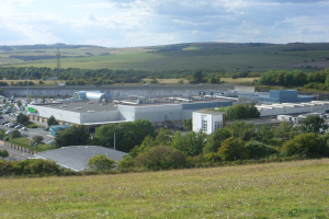 Eine große Fabrik in einem Feld umgeben von Gebäuden, Bäumen, Pflanzen, Gras, Fahrzeugen, Masten und einem bewölkten Himmel.