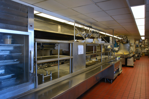 Gewerbeküche mit Edelstahl-Theke, elektrischer Deckenbeleuchtung, hängenden Utensilien, einem Spülbecken, Wasserhähnen, Schränken und Gegenständen auf Regalen.