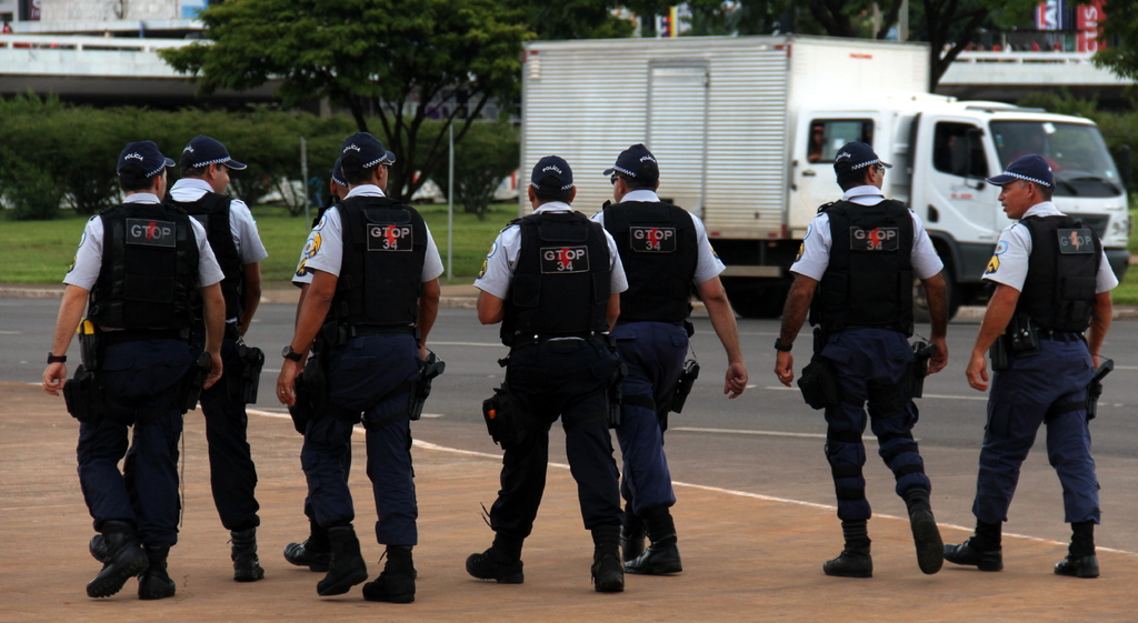 Eine Gruppe von Polizisten in Uniform, einschließlich Mützen, Hemden, kugelsicheren Westen, Hosen und Schuhen, die auf einer Straße mit einem geparkten Lkw auf der rechten Seite neben einem Zaun, Gebäuden, Bäumen, Pflanzen, Pfählen und Bannern im Hintergrund und einer Brücke oben rechts gehen.