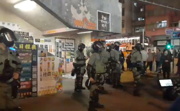Polizisten in Schutzausrüstung stehen vor einer Menge von Helmet tragenden Demonstranten mit Waffen, mit Plakaten an Wänden, Deckenleuchten und Gebäuden im Hintergrund, einschließlich einer Verkehrsampel mit einer Texttafel in Hong Kong.