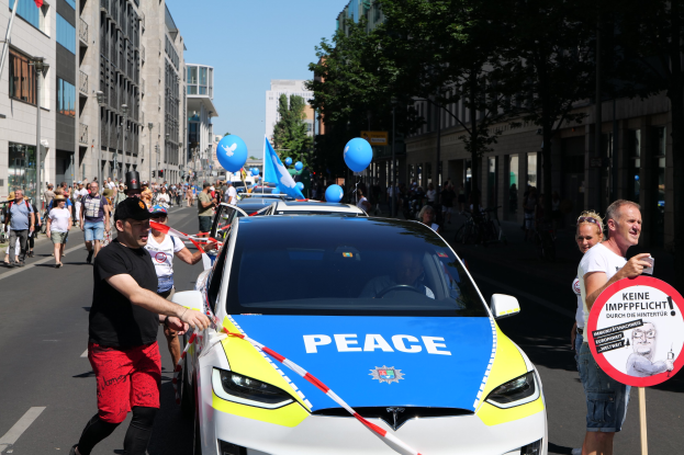 Eine Gruppe von Menschen geht neben einem Polizeiwagen her, der mit blauen, gelben und roten Schleifen geschmückt ist und auf dem das Wort "Frieden" zu sehen ist, vor einer Kulisse aus Gebäuden, Bäumen und Fahnen unter einem klaren blauen Himmel.