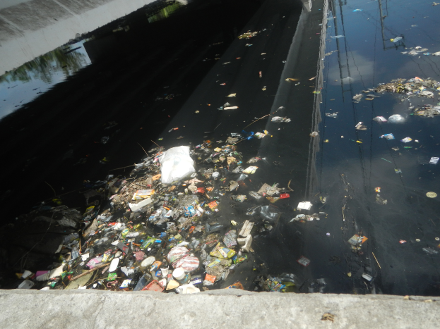 Ein Fluss voller Müll neben einer Brücke, mit einer Wand auf der linken Seite und einer Straße unten.