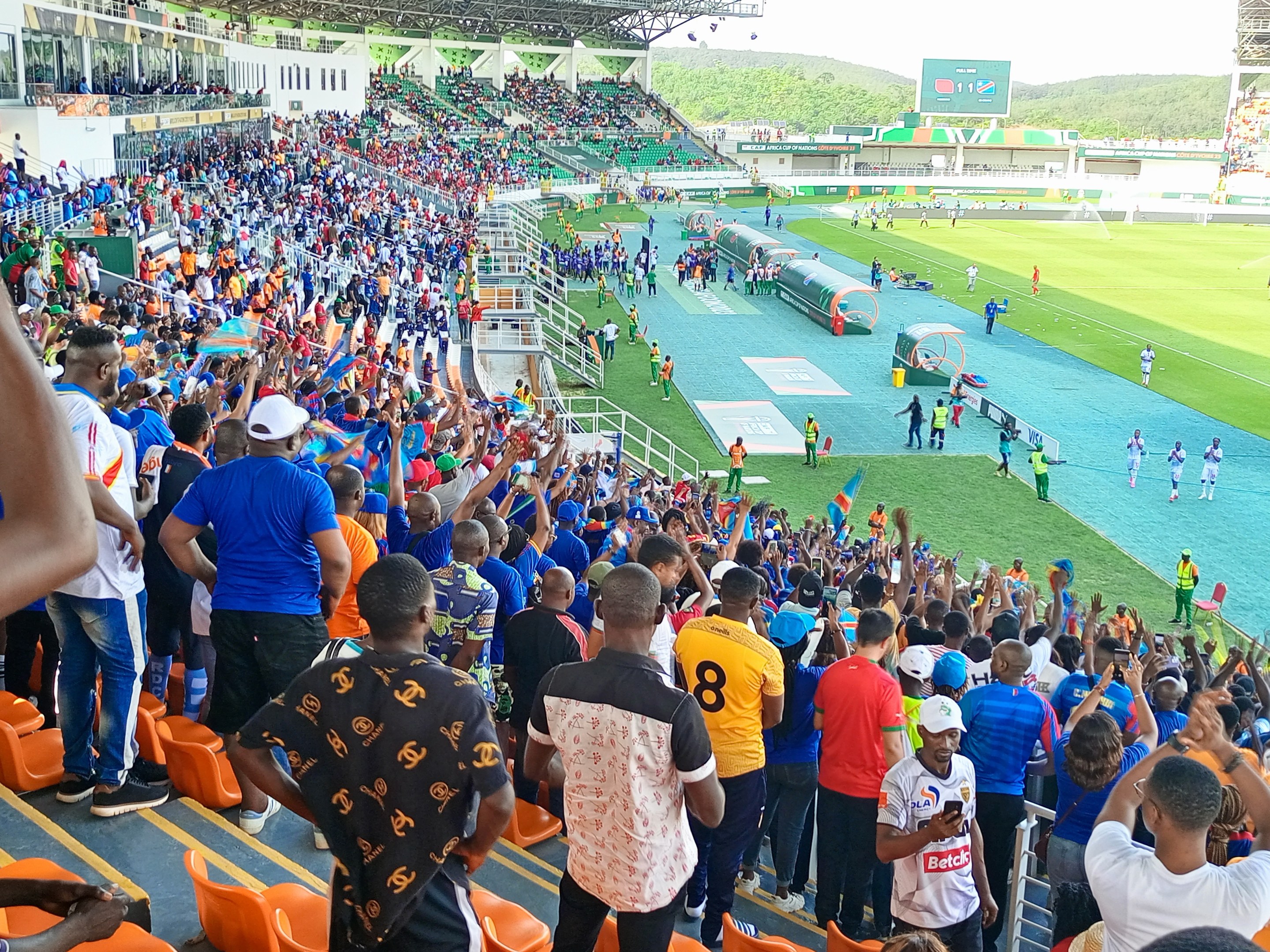 Eine große Menschenmenge versammelt sich vor einem eingezäunten Fußballfeld, einige sitzen auf Stühlen, andere stehen, unter einem klaren blauen Himmel mit Hügeln im Hintergrund und warten auf das Spiel der 2019 African Cup of Nations.