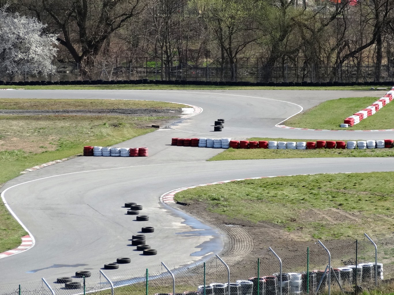Rennstrecke mit zahlreichen Reifen entlang ihrer Ränder, umgeben von Gras und Maschendrahtzaun, mit Bäumen im Hintergrund.