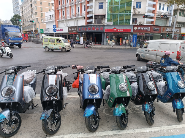 Eine Reihe von elektrischen Rollern, die auf einer Straße geparkt sind, mit einer Person in einer Mütze davor, vor einem Hintergrund aus Gebäuden, Verkehrsampeln, Bäumen und einem klaren blauen Himmel.