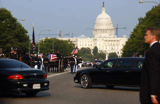 Eine Trauerprozession vor dem Kapitolsgebäude, mit vielen Menschen in Mützen und mit Fahnen, Fahrzeugen auf der Straße, Laternenpfählen, Bäumen und Gebäuden mit Fenstern im Hintergrund, unter einem klaren Himmel.