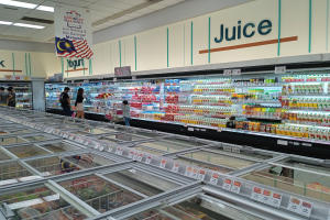 Ein hell erleuchteter Saftladen in einem Supermarkt zeigt Flaschengetränke und frisches Obst auf Regalen, mit einigen Kunden, die stöbern und Deckenleuchten, die die Gegend beleuchten.